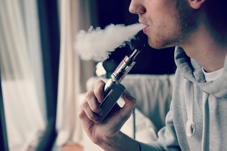 Young man uses a vaping device, puffing out a cloud of smoke.