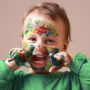 Humorous photo of cute cheerful child girl showing her hands and face painted in bright colors.