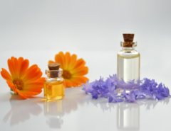 Glass bottles of liquid with cork stoppers displayed beside orange and purple flowers on a white background.