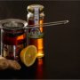 A jar of golden honey, a metal tea infuser resting across a glass of dark amber tea, a half lemon, and a piece of fresh ginger root arranged on a dark tabletop.