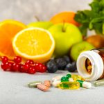 Various fruits and leafy vegetables scattered across a countertop with an open bottle with various pills and vitamins spilling out in front.