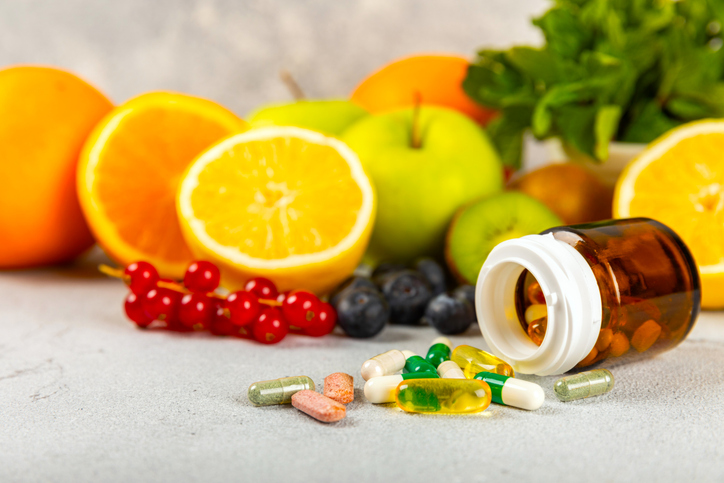 Various fruits and leafy vegetables scattered across a countertop with an open bottle with various pills and vitamins spilling out in front.